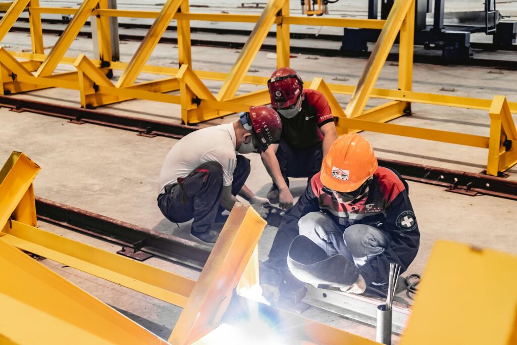 Three construction workers welding metal structures indoors, wearing safety gear.